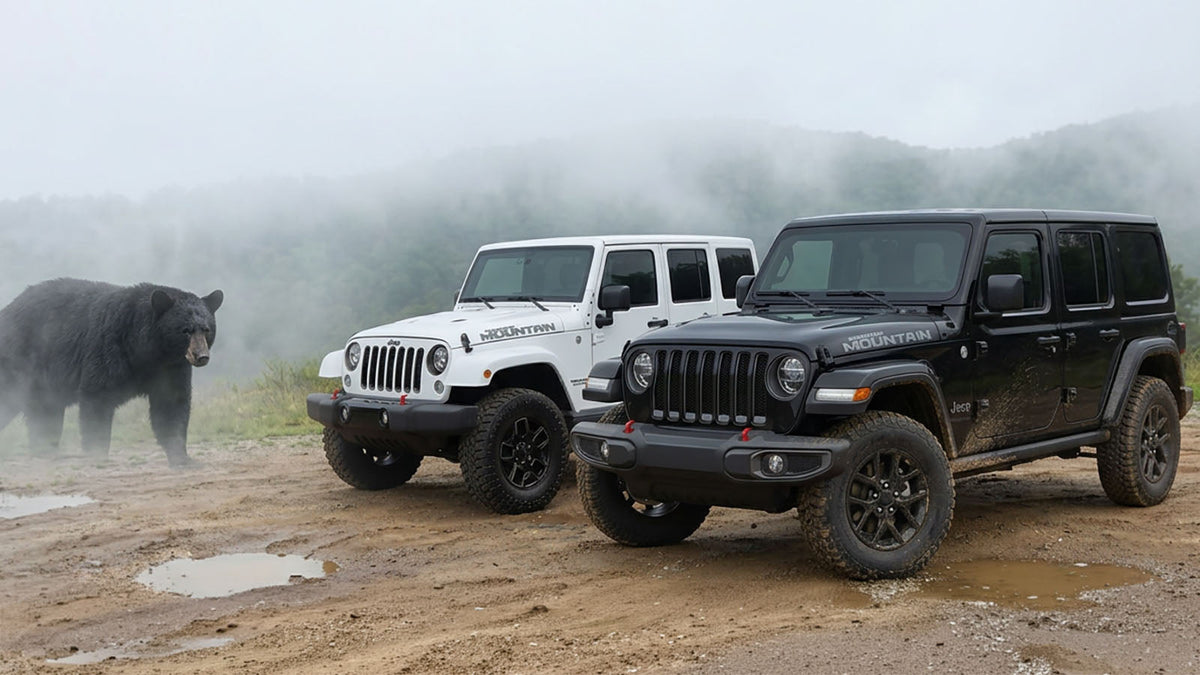 A white 2017 Jeep Wrangler JK Smoky Mountain Edition and a black 2027 Wrangler JL Smoky Mountain parked on a wet gravel trail, both wearing Smoky Mountain hood decals, with a black bear standing in the fog behind them