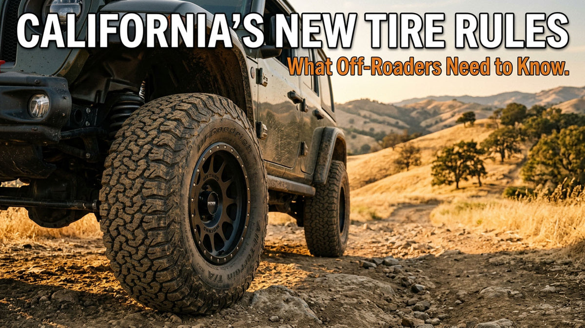 A close-up of a BFGoodrich All-Terrain tire on a Jeep Wrangler parked on a dirt trail with golden California hills in the background