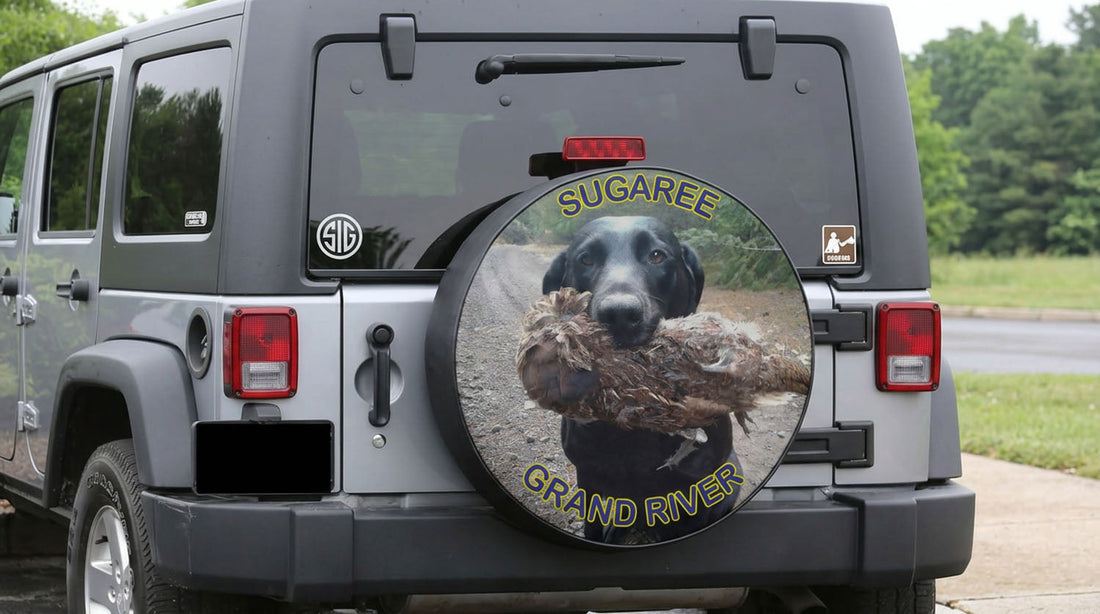Custom spare tire cover on a silver Jeep Wrangler showing a black Labrador retriever carrying a game bird, personalized with the names Sugaree and Grand River