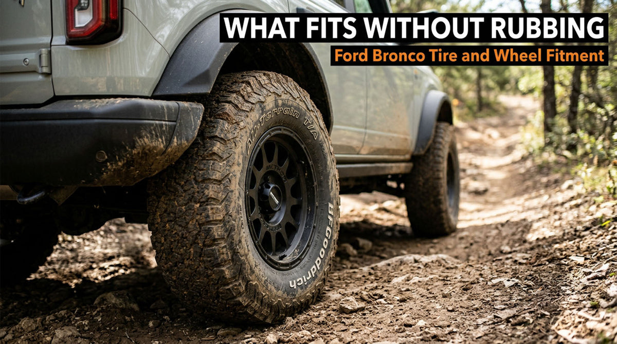Close-up of the rear wheel of a light gray Ford Bronco on a dirt trail, showing a mud-caked BFGoodrich all-terrain tire on a bronze wheel under a flared fender, with a headline reading What Fits Without Rubbing