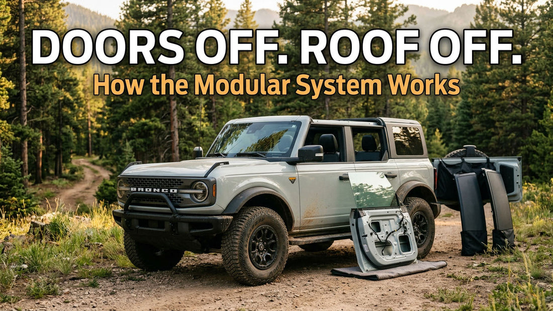 A light gray four-door Ford Bronco parked on a dirt forest trail with the front passenger door removed and resting on a mat, and roof panels and soft top pieces stacked against the rear of the vehicle