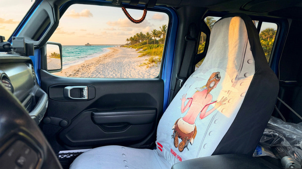 Ready for Takeoff pin-up aviator seat cover installed on the passenger seat of a blue Jeep parked at the beach at sunset