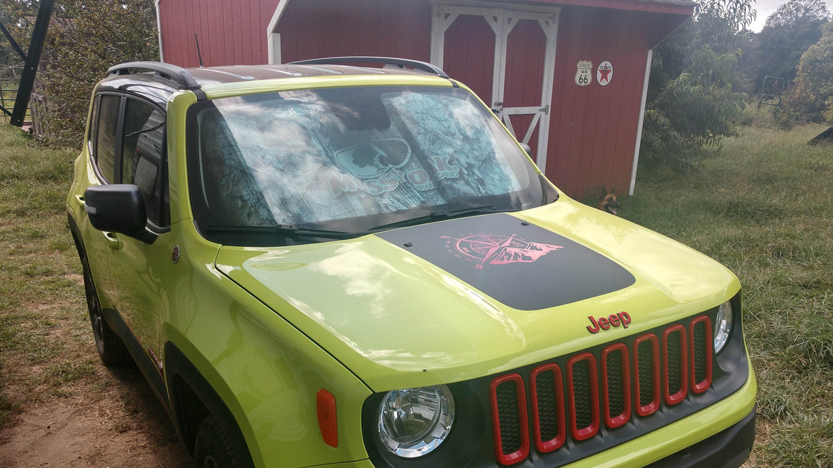 Bright green Jeep Renegade with a matte black hood decal featuring a pink compass and mountain design, parked on grass beside a red barn