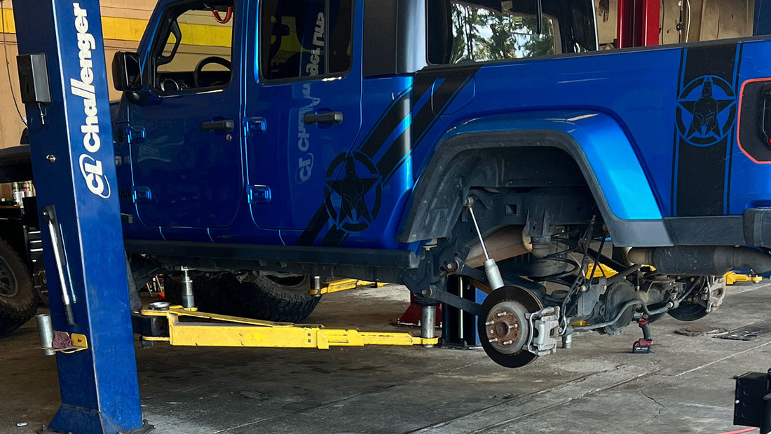 Blue Jeep Gladiator raised on a two-post shop lift with the rear wheel removed, exposing the five-lug wheel hub, brake rotor and caliper