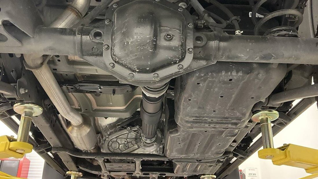 Underside view of a Jeep Wrangler on a shop lift showing the rear differential cover, axle tubes, and driveshaft