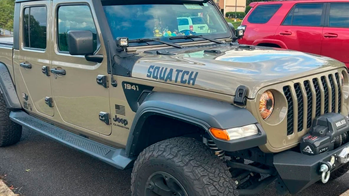 A tan Jeep Gladiator with a SQUATCH hood decal, American flag graphic, and front winch parked in a lot