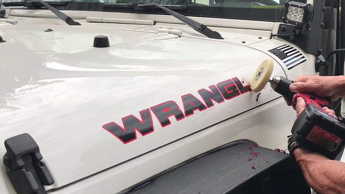 Hands warming an old vinyl decal with a hair dryer and peeling it off a Jeep body panel with a plastic scraper, leftover adhesive residue visible on the paint