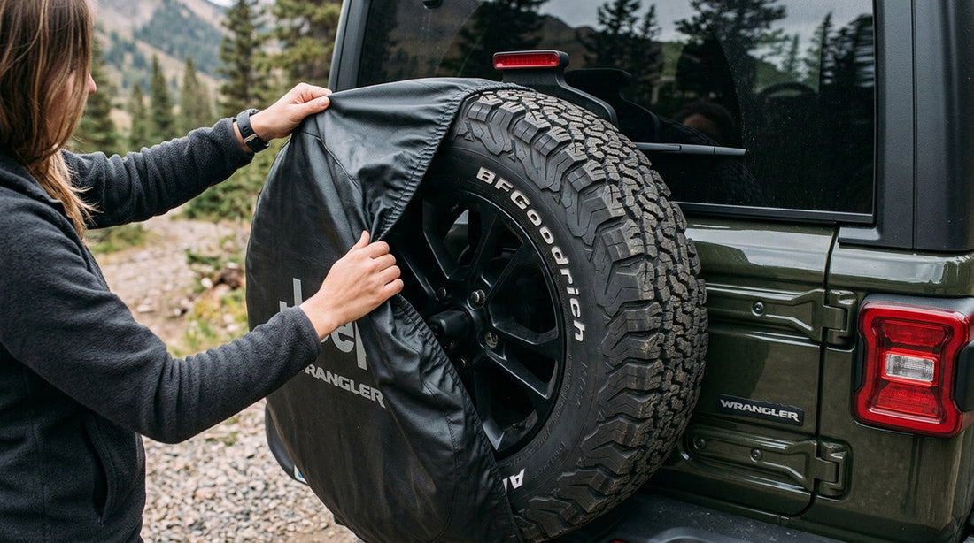 Hands easing a black fabric spare tire cover partway off the rear-mounted spare wheel of an olive green Jeep Wrangler on a mountain trail
