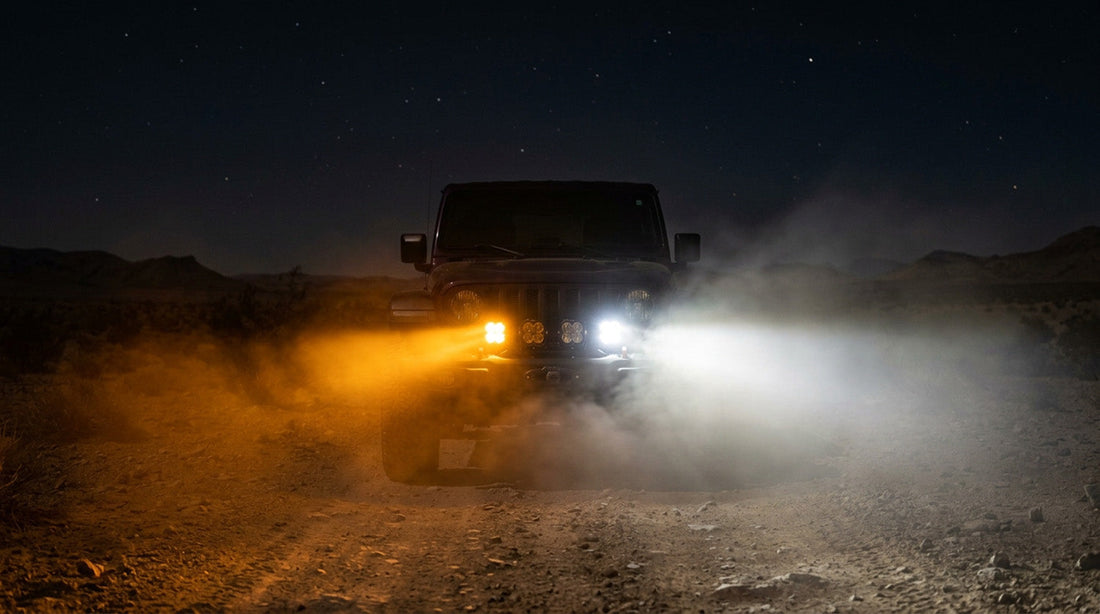 Amber and white off-road pod lights on a Jeep at night, amber cutting through dust while white scatters into glare