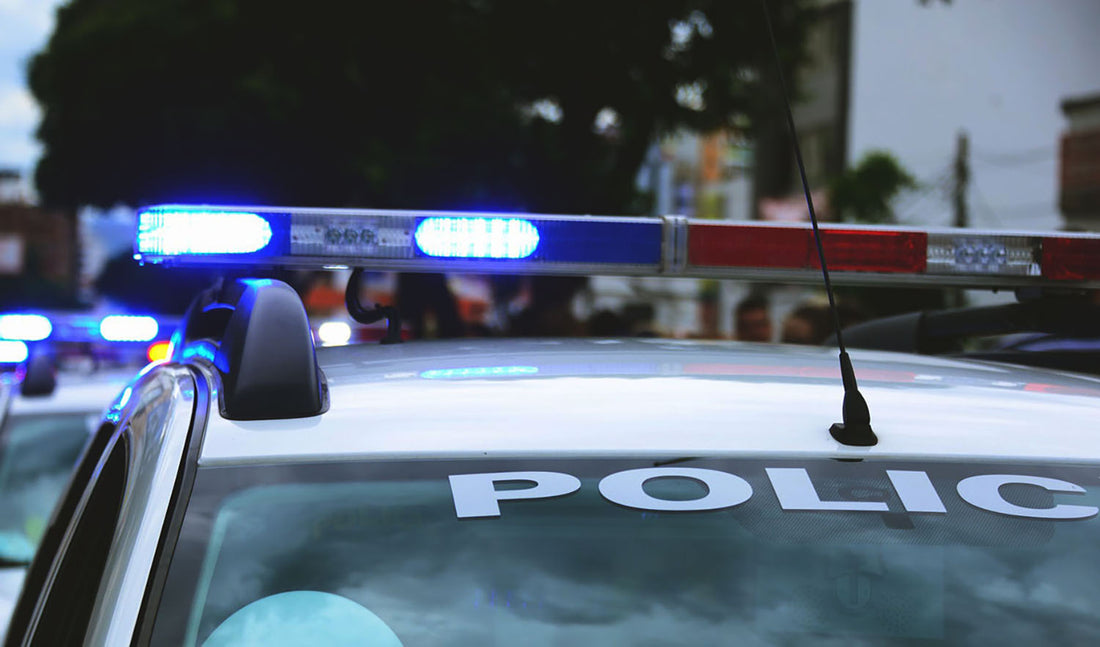 Police cruiser light bar with blue and red lights lit, POLICE lettering across the windshield, with a line of vehicles and spectators blurred behind it, the escort at the head of a Back the Blue Jeep rally