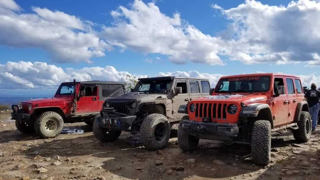 Three Jeep Wranglers, two red and one tan, parked side by side on a rocky mountain overlook under a blue sky with clouds