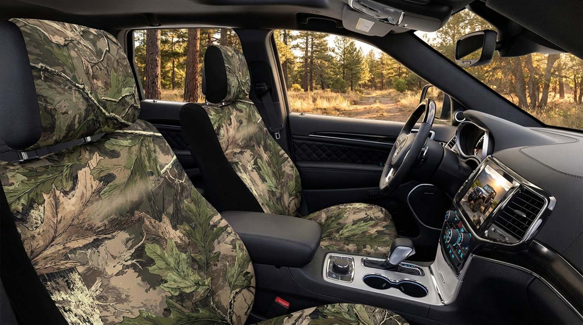 Oakbrush-style woodland camo seat covers on the front seats of a Jeep Grand Cherokee, parked on a dirt road through fall pine woods