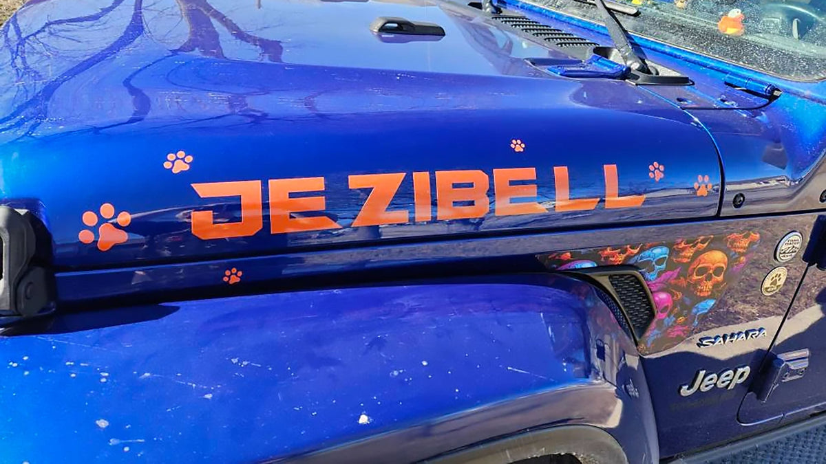 Orange custom hood lettering reading JEZIBELL with matching paw print decals on the hood of a blue Jeep Wrangler JL Sahara, with a colorful skull fender vent decal alongside