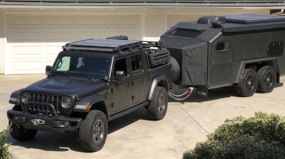 Blacked-out Jeep Gladiator with a solar panel roof rack, light bar and traction boards, towing a dark tandem-axle off-road overland trailer on a driveway