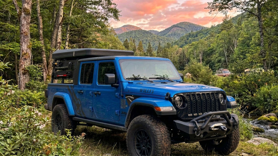 Blue Jeep Gladiator Willys with a bed rack, rooftop tent and front winch bumper parked beside a mountain creek at sunset, with forested ridges in the background