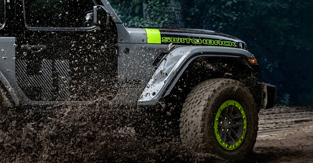 2027 Jeep Wrangler Switchback in Sting-Gray with a contrasting black door, Gecko green Switchback hood graphic, and Gecko beadlock wheel ring, throwing mud on a forest trail