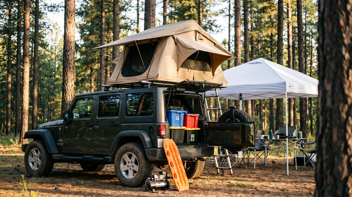 Jeep Wrangler Unlimited at a pine forest campsite with a soft shell rooftop tent on crossbars, a white pop-up canopy over a camp table, colored storage bins in the cargo area, and orange traction boards and an air compressor by the rear tire