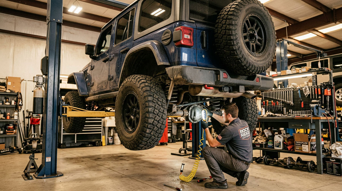 Technician kneeling under a blue Jeep Wrangler JL Unlimited raised on a two-post shop lift, working on the driveline with an impact tool, spare tire mounted on the tailgate