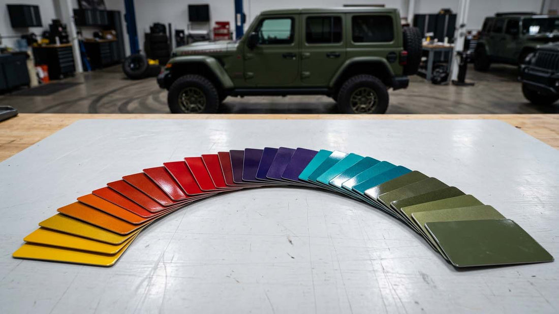 Automotive paint sample chips in Jeep factory colors fanned out in an arc on a workbench, with a Sarge Green Jeep Wrangler in the shop behind, illustrating Jeep Wrangler paint colors by year
