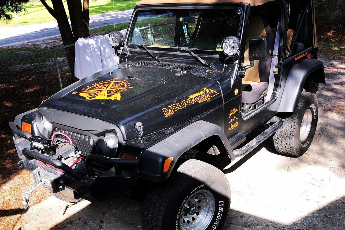 Black 1997 Jeep Wrangler TJ with the doors off, a winch bumper, KC driving lights, and a mountain compass hood graphic, parked on a shaded driveway