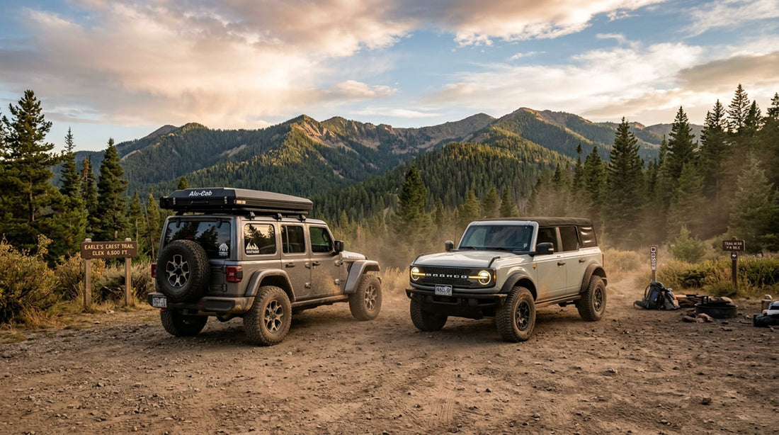 Jeep Wrangler JL Unlimited with a rooftop tent and tailgate spare parked beside a four-door Ford Bronco at a dirt trailhead staging area at sunset