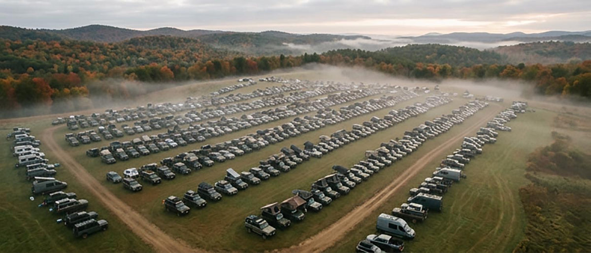 Aerial view of hundreds of 4x4s and camper vans parked in rows with rooftop tents open across an open grass camping field at dawn, mist over the field and autumn foothills behind