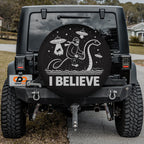 the back of a black Jeep Wrangler with a large, round spare tire cover on the rear of the vehicle. The cover features a cartoon image of a monkey riding a dinosaur on a boat, along with the text "I BELIEVE".