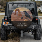 Arches National Park spare tire cover on a Jeep Wrangler, showing a raven on a sandstone ledge beside Delicate Arch