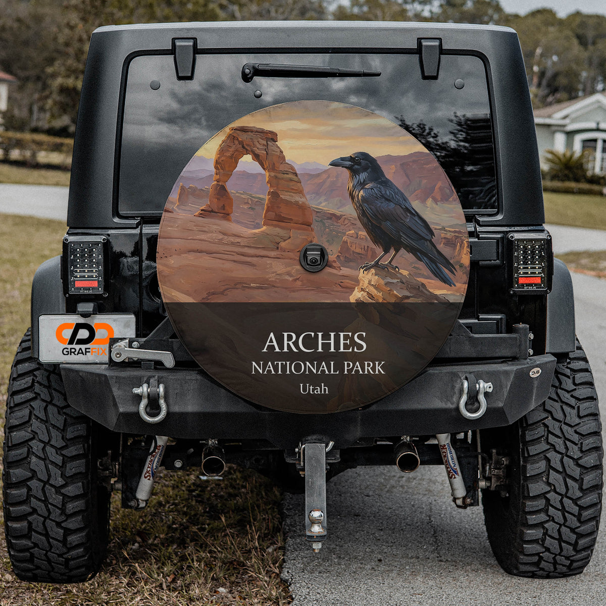 Arches National Park spare tire cover on a Jeep Wrangler, showing a raven on a sandstone ledge beside Delicate Arch