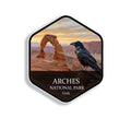 Hexagonal Arches National Park sticker badge with a common raven perched on a sandstone fin above red rock arches at sunrise