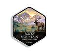 Hexagonal Rocky Mountain National Park sticker badge with a bull elk on a rocky rise above an alpine meadow below snow-streaked peaks at sunrise