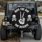 the back of a black Jeep Wrangler with a large black wheel cover featuring a skull and crossbones design and the text "Have the day you deserve".