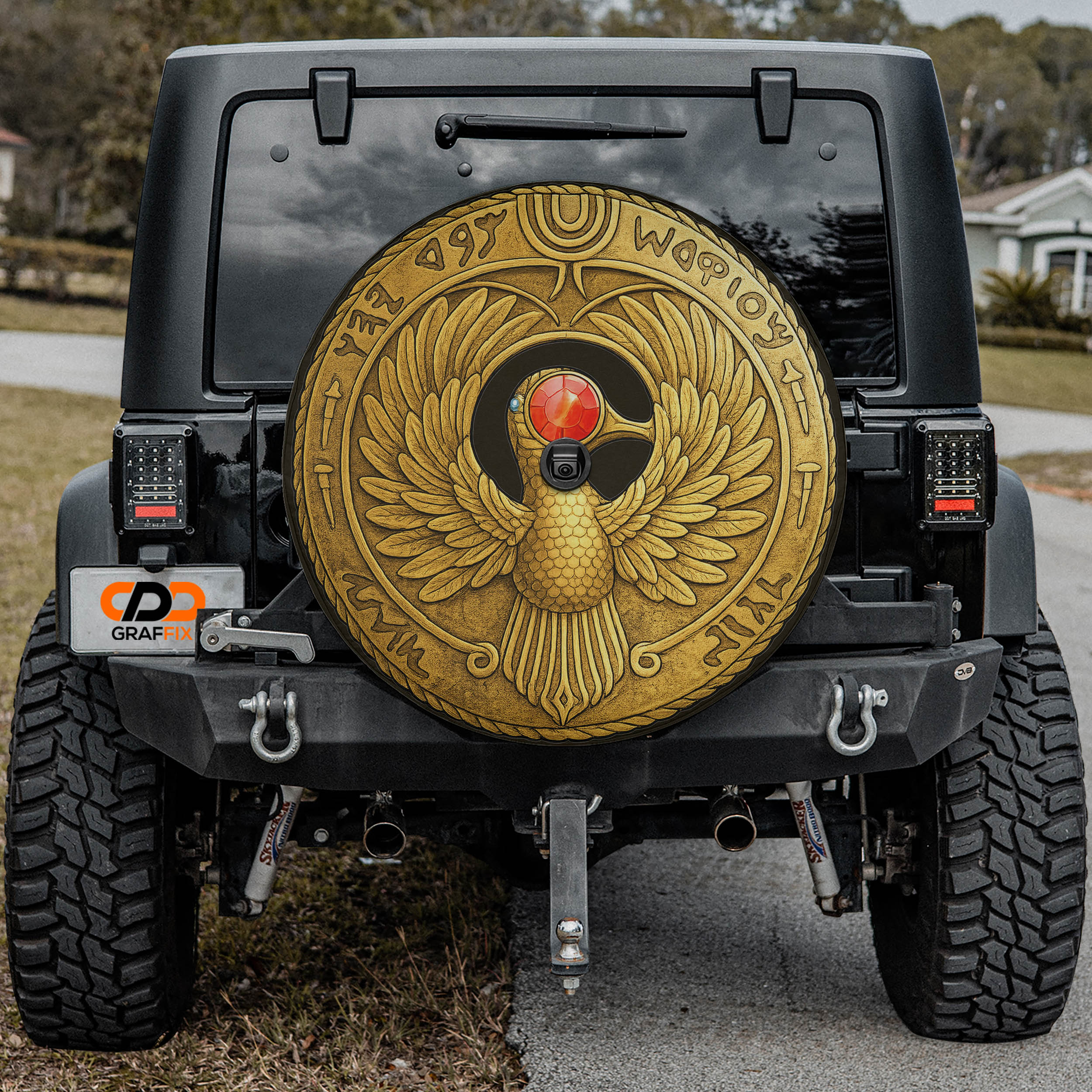 a black Jeep Wrangler with a large, ornate gold-colored wheel cover featuring an Egyptian-style bird design.