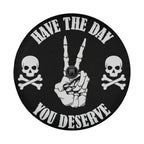 a black and white design with a hand making a peace sign and two skull and crossbones symbols on either side. The text "HAVE THE DAY YOU DESERVE" is displayed around the design.